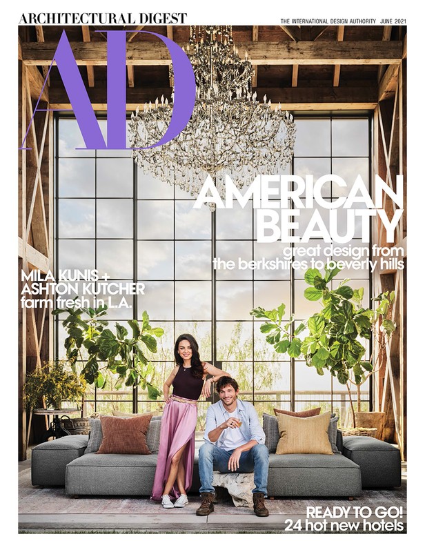Mila Kunis and Ashton Kutcher sit on a grey sofa in front of a large window in Architectural Digest magazine cover, plants are placed in front of the window.