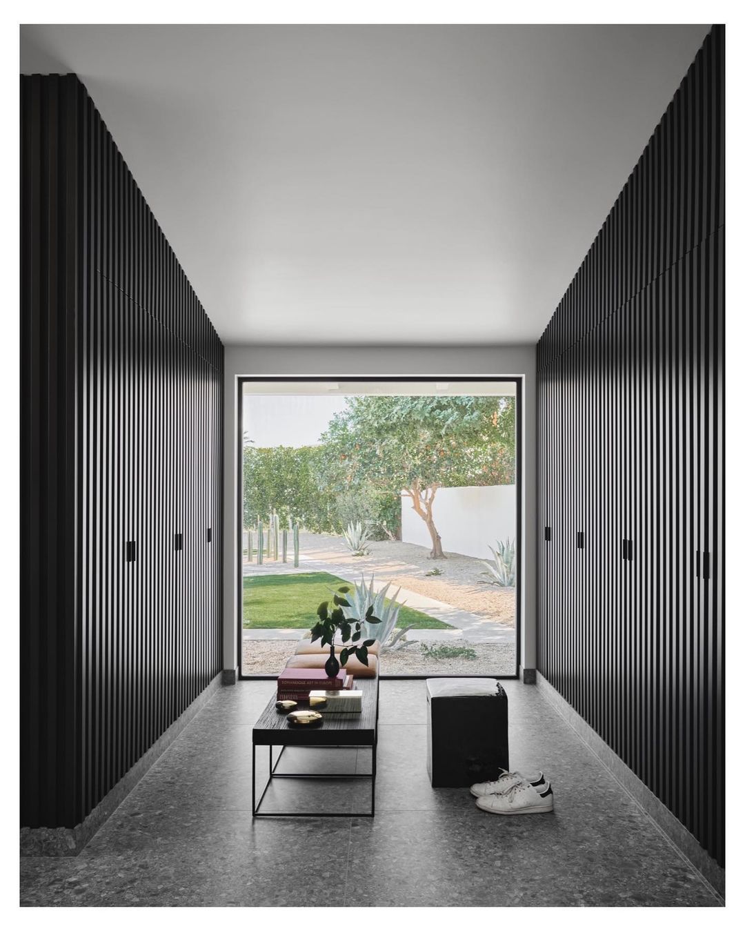 Hallway view features black wood slat walls, terrazzo floor, modern black table with objects, and a view to a green garden.