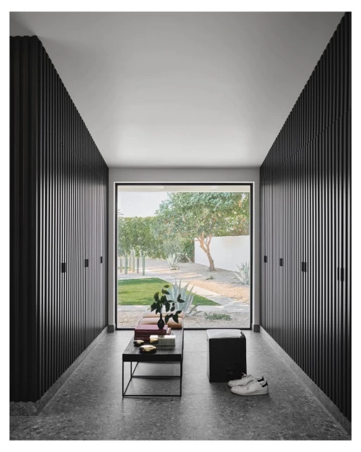 Hallway view features black wood slat walls, terrazzo floor, modern black table with objects, and a view to a green garden.