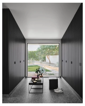 Hallway view features black wood slat walls, terrazzo floor, modern black table with objects, and a view to a green garden.