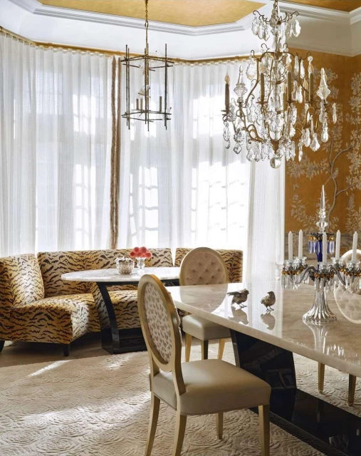 A well-lit dining room features tan upholstered chairs, banquette seating with tiger print, and crystal chandeliers in a home interior.