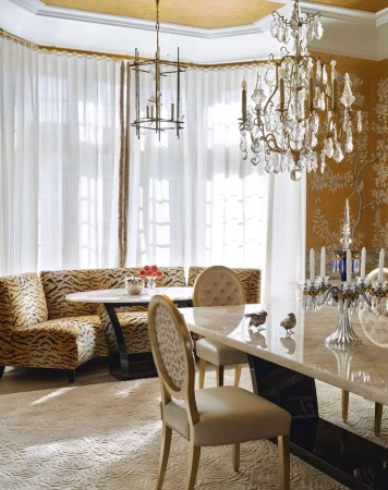 A well-lit dining room features tan upholstered chairs, banquette seating with tiger print, and crystal chandeliers in a home interior.