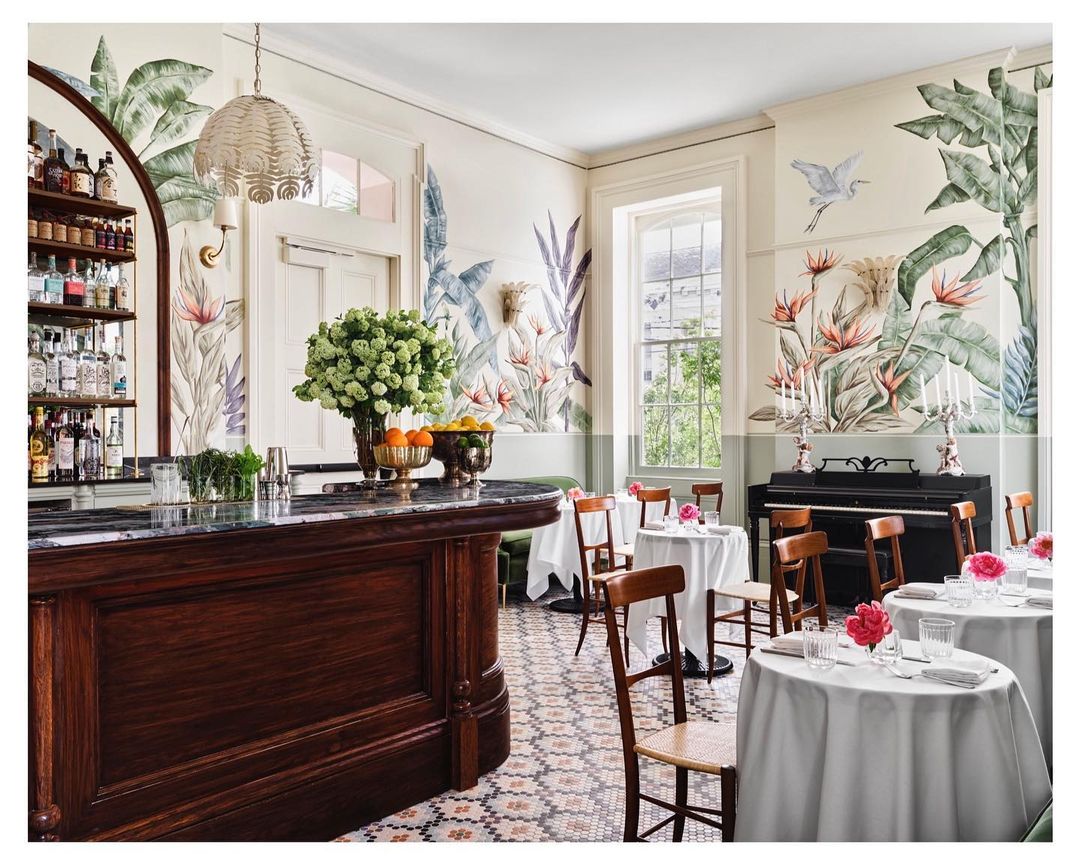 Interior of a dining room featuring tropical wallpaper, wood furniture, circular tables with white tablecloths, flowers and bar area.