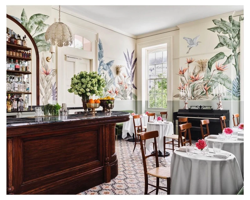 Interior of a dining room featuring tropical wallpaper, wood furniture, circular tables with white tablecloths, flowers and bar area.