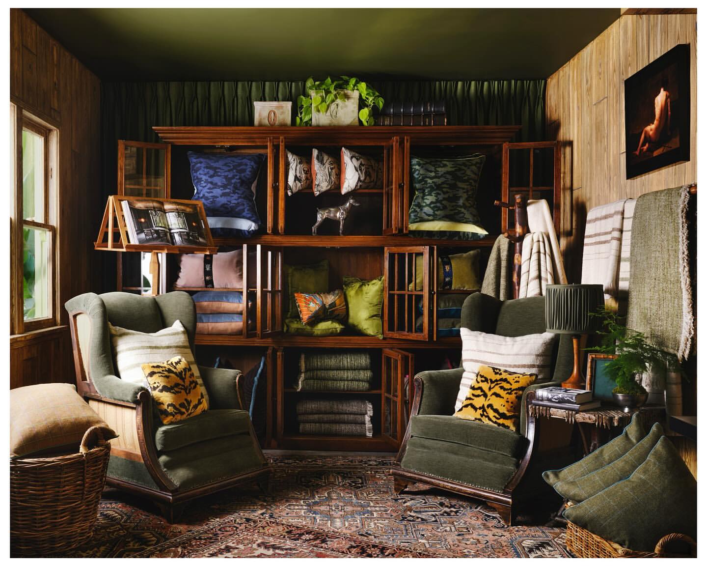 A warm and inviting living room features two green wingback chairs, a wooden cabinet filled with pillows and throws, and patterned rug.
