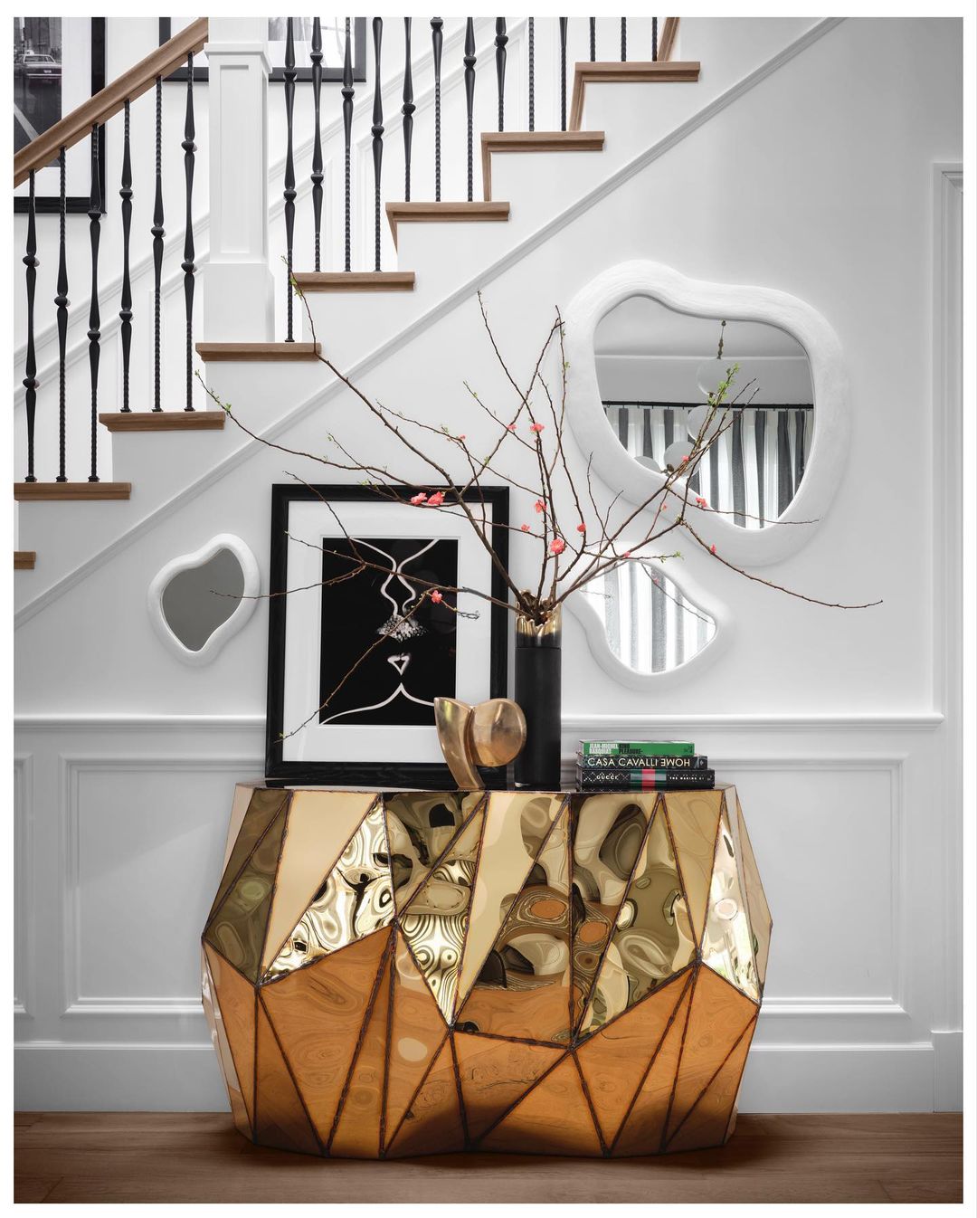 Interior design featuring a geometric gold console table, artwork, mirrors, and staircase with a white wall background in a home.