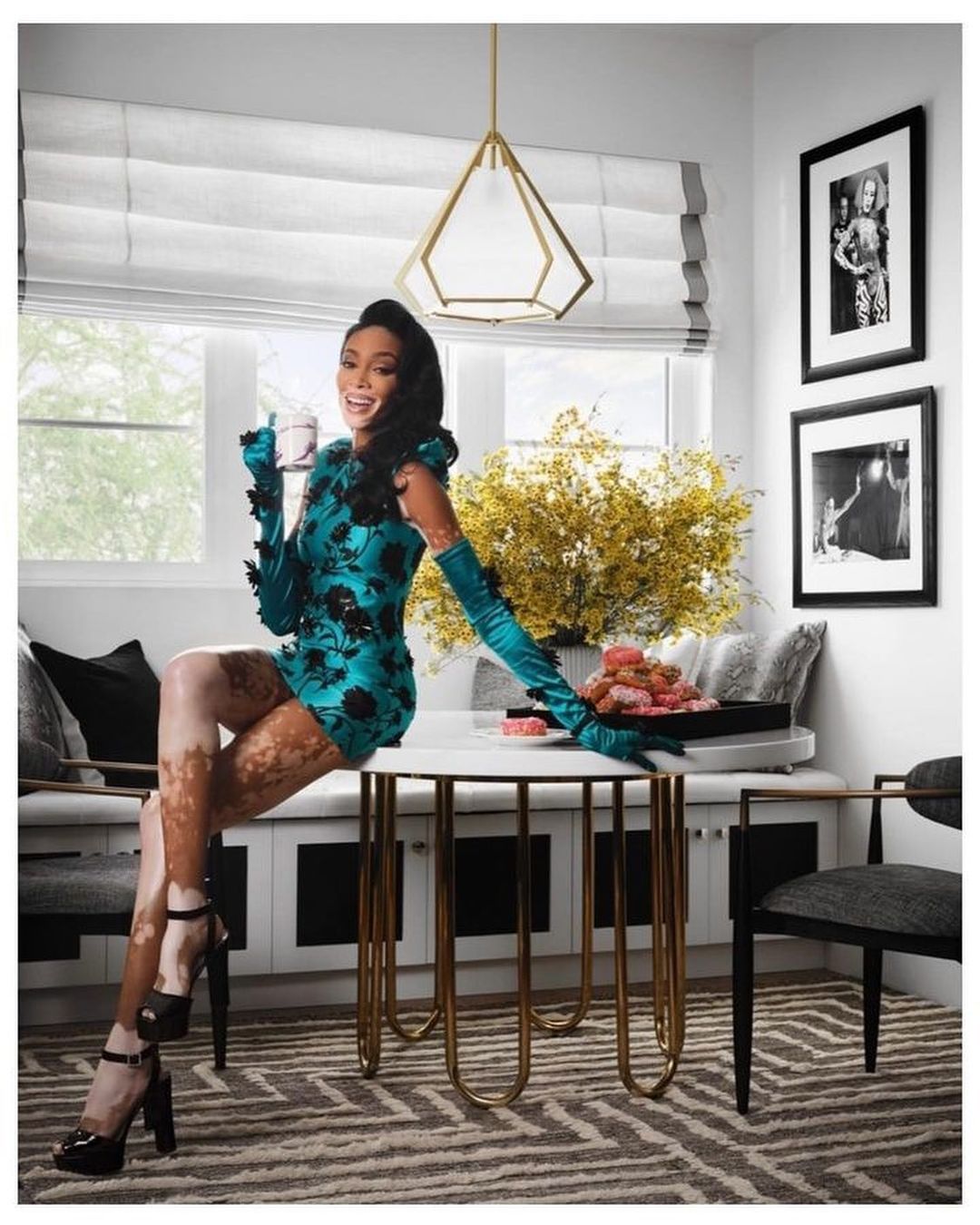 Model Winnie Harlow in a green floral mini dress and gloves, sitting on a table in an indoor setting with donuts.
