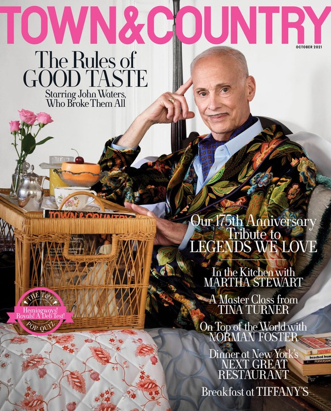Magazine cover featuring John Waters in a floral jacket, seated with a wicker tray holding flowers, dessert, and book, October 2021.