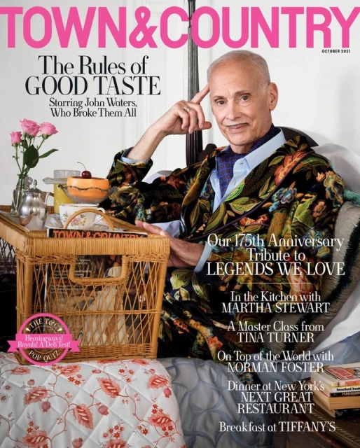 Magazine cover featuring John Waters in a floral jacket, seated with a wicker tray holding flowers, dessert, and book, October 2021.