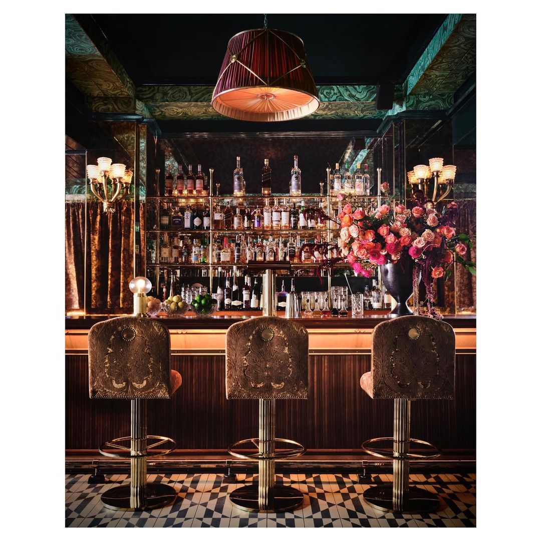 Elegant interior featuring a bar with three stools, a well-stocked liquor shelf, and a flower arrangement under a decorative lamp.