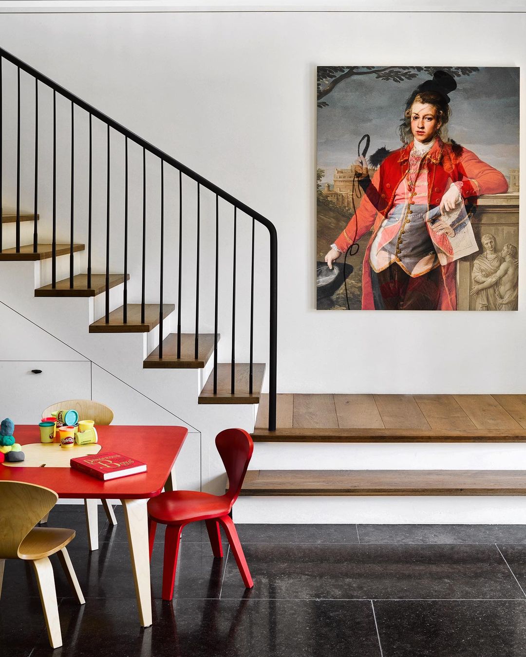 Interior shot of a modern home featuring a painting of a man, a black staircase, and a red table with chairs in a cozy space.