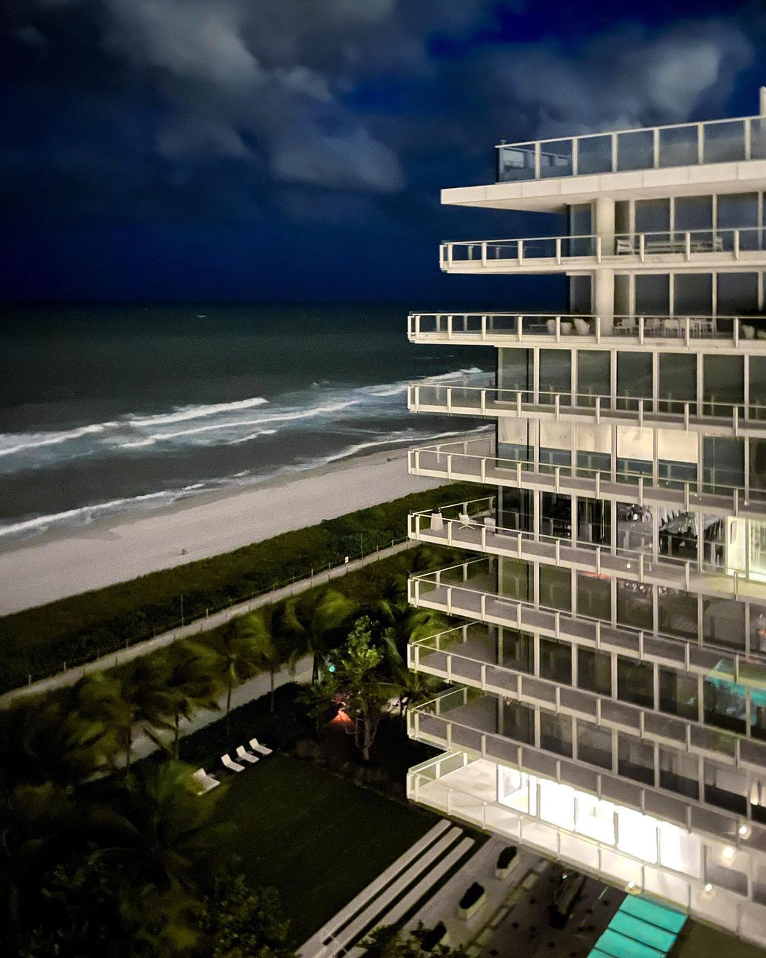 Night view of a modern high-rise building overlooking a beach with breaking waves under a cloudy sky, captured from an elevated angle.