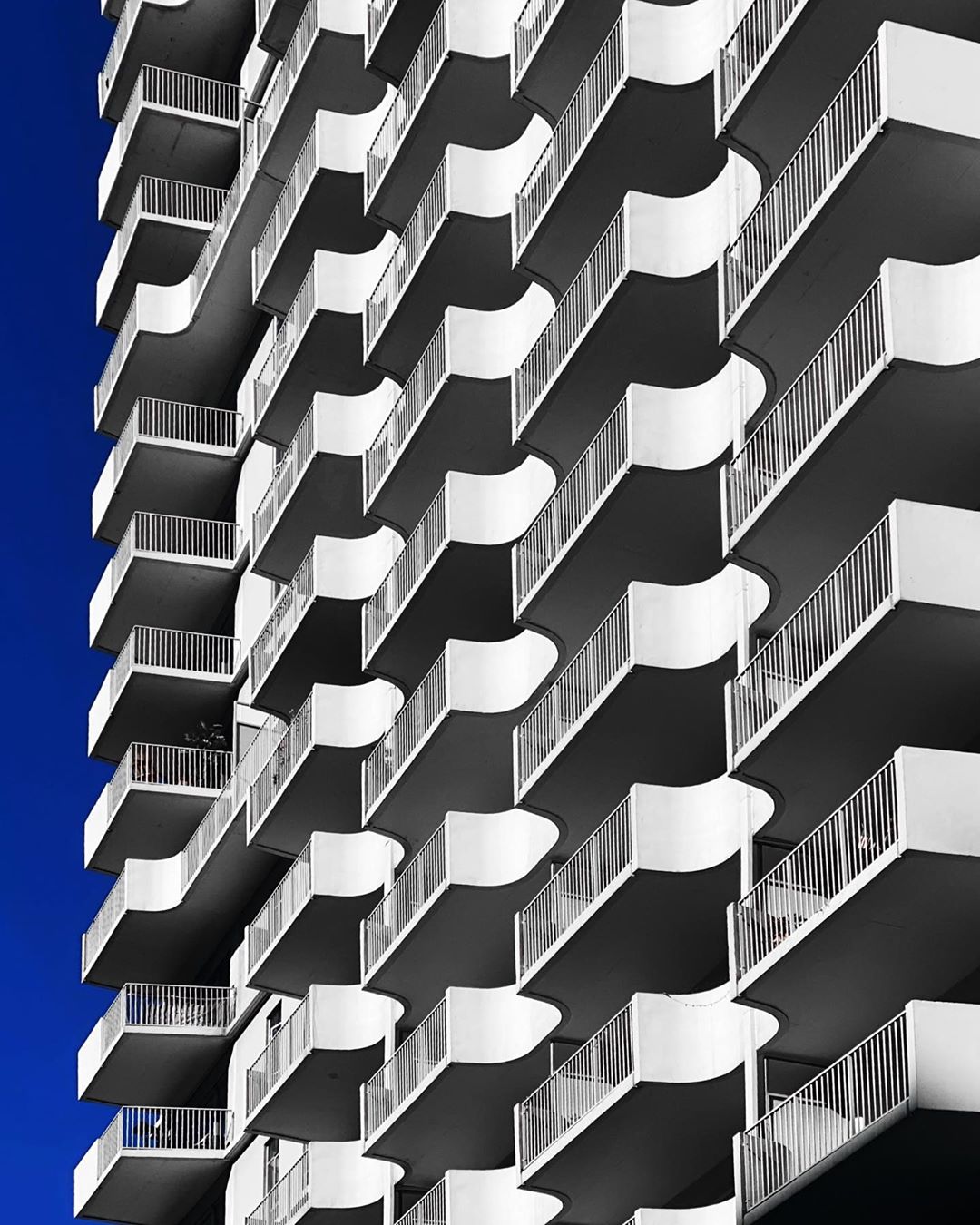 Architectural detail of a tall modern building with rows of balconies against a vivid blue sky, showcasing geometric patterns.