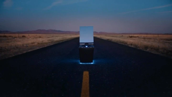 Modern dark object with reflective screen on highway at night. Desert landscape under twilight sky with blue accent lighting.