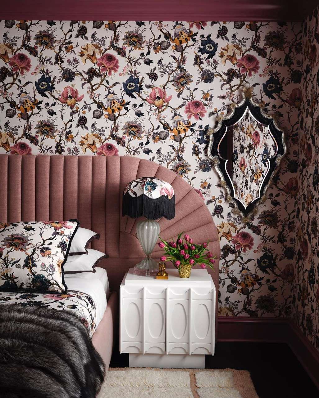 Interior bedroom with floral wallpaper, a pink headboard, white nightstand, lamp, tulips, pillows, and a decorative mirror on the wall.