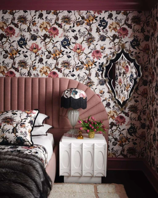 Interior bedroom with floral wallpaper, a pink headboard, white nightstand, lamp, tulips, pillows, and a decorative mirror on the wall.