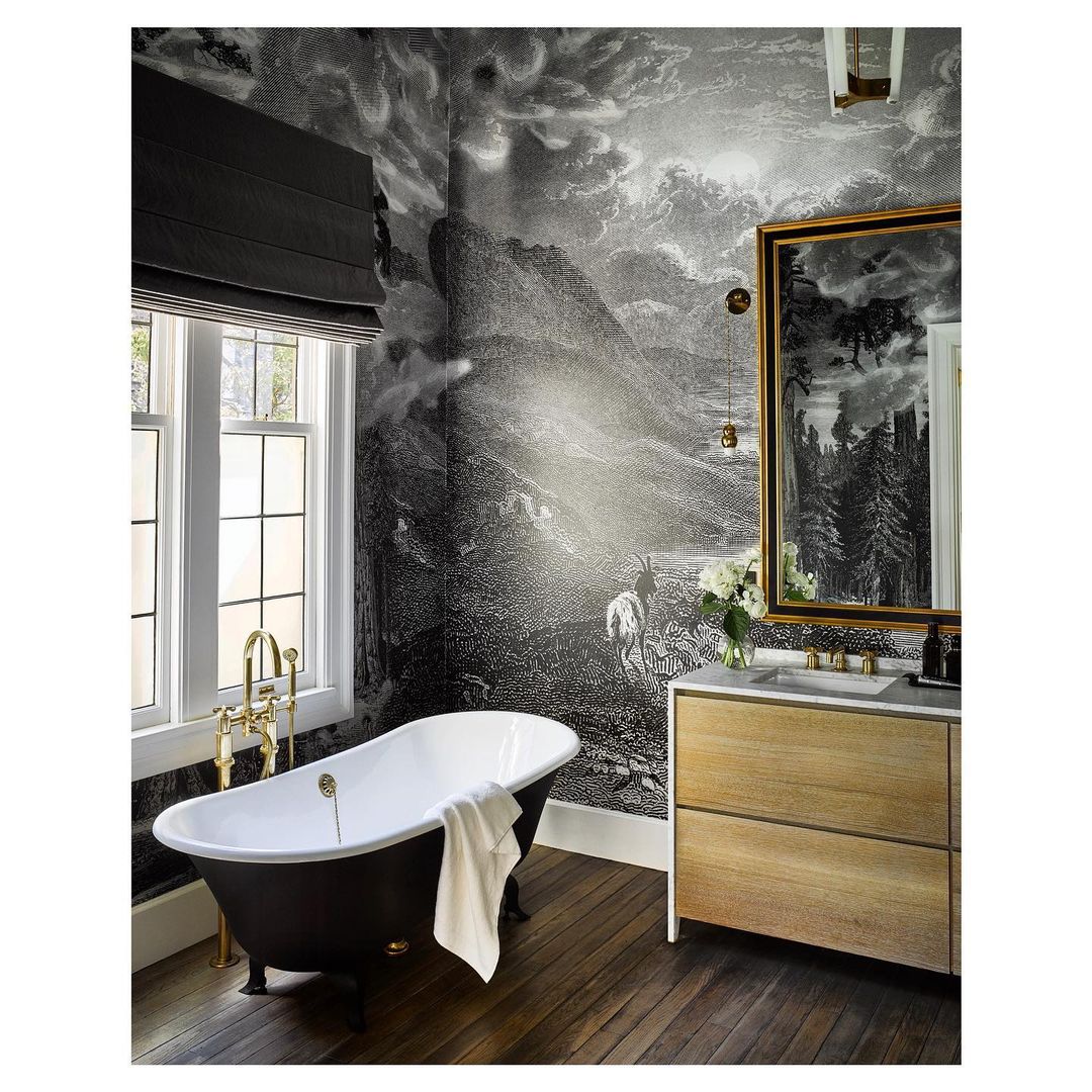 Bathroom interior featuring a clawfoot tub with gold fixtures, a wooden vanity, and a decorative landscape-themed wallpaper and gold-framed mirror.