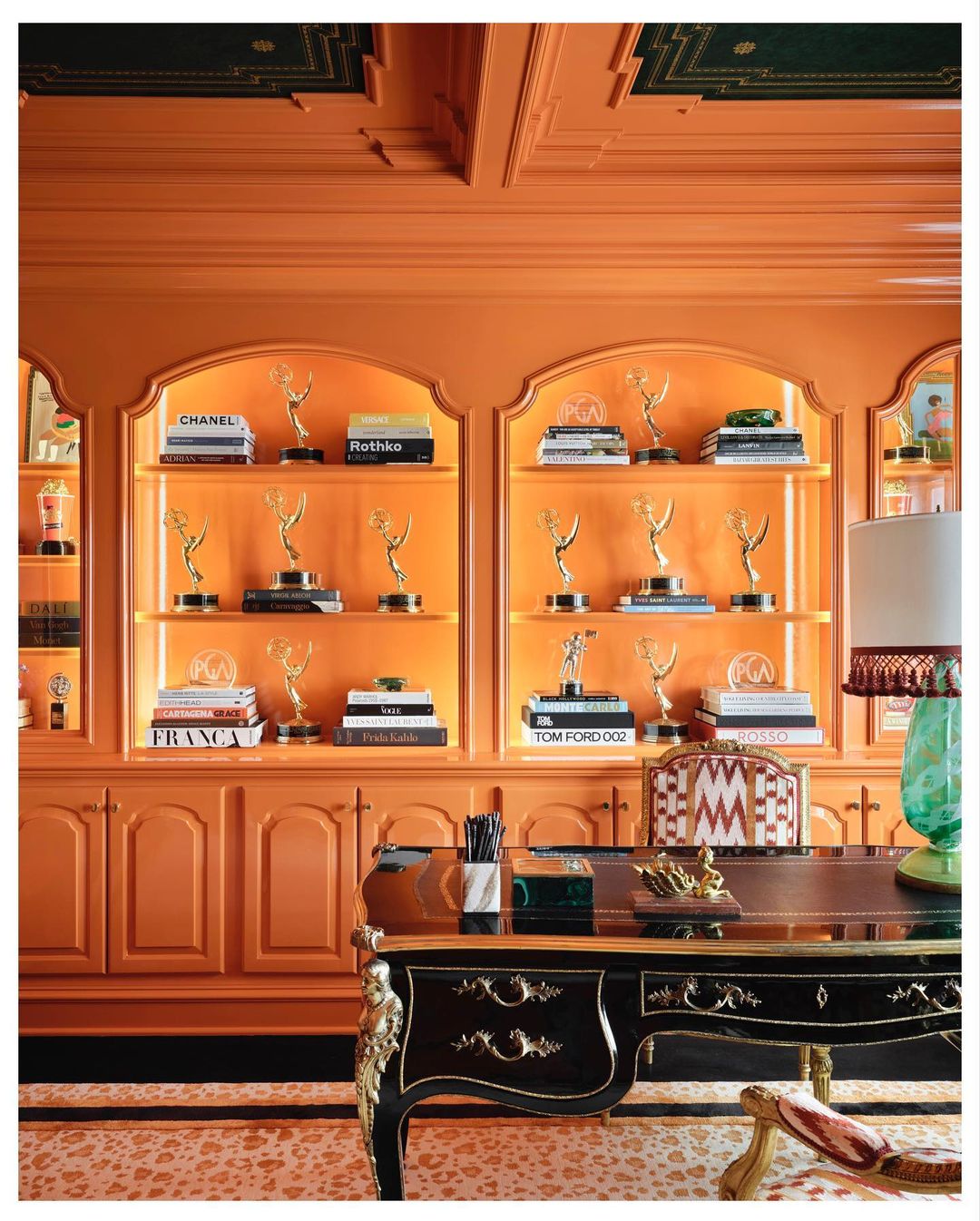Orangey-toned home office interior featuring a black desk, built-in bookcase lined with books, and Emmy awards trophies on shelves.