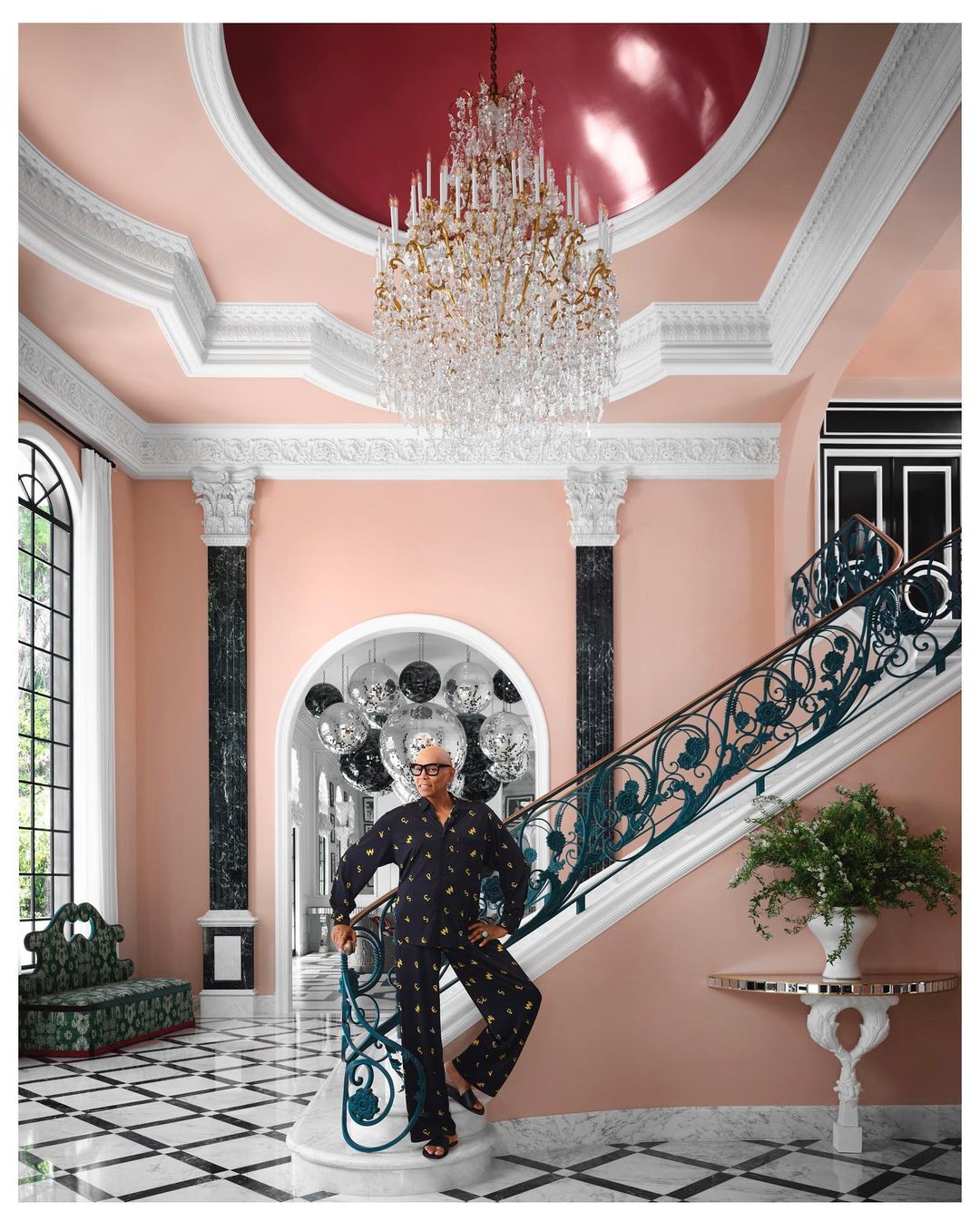 Elegant woman in patterned pajamas poses on the staircase of a grand foyer with chandelier and checkered marble flooring.