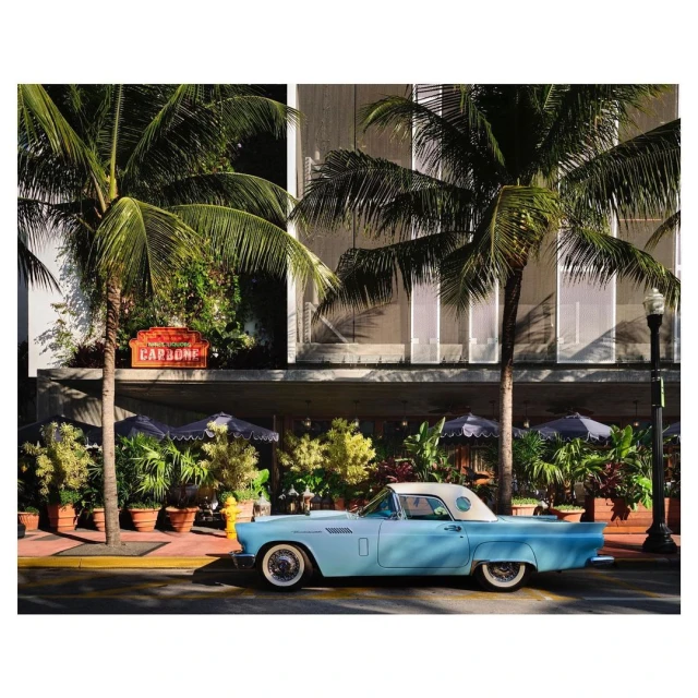 Turquoise vintage car parked along a street with palm trees and lush greenery in front of a building with awnings and umbrellas.