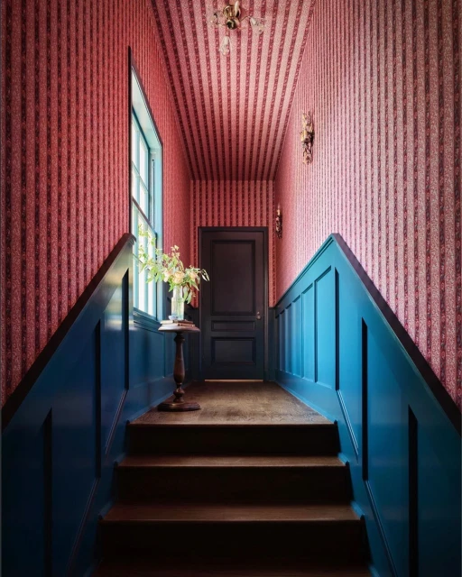 Hallway interior featuring blue wainscoting and red patterned wallpaper, with a small table holding flowers and a dark wood staircase.
