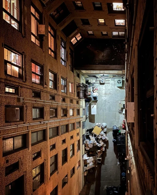 Urban alleyway view from above with brick buildings, many windows, people working, trash cans, and light reflecting on wet pavement.