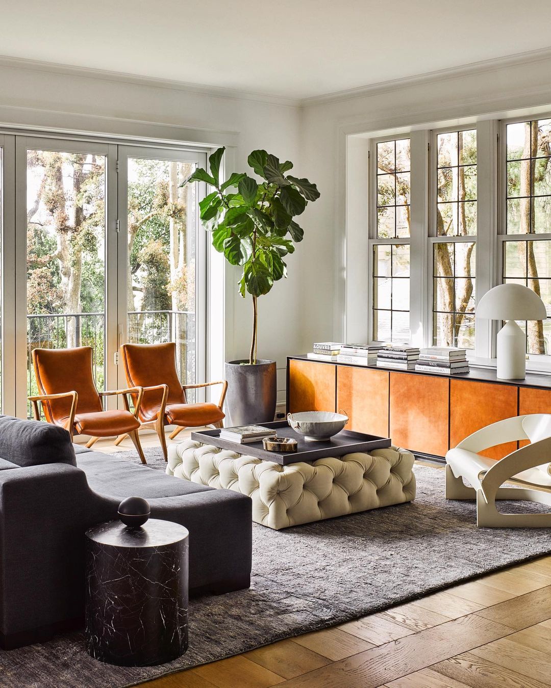 Living room interior with a grey sofa, tufted ottoman, and wooden floors, bathed in natural light streaming through large windows.