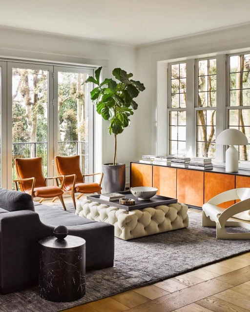 Living room interior with a grey sofa, tufted ottoman, and wooden floors, bathed in natural light streaming through large windows.
