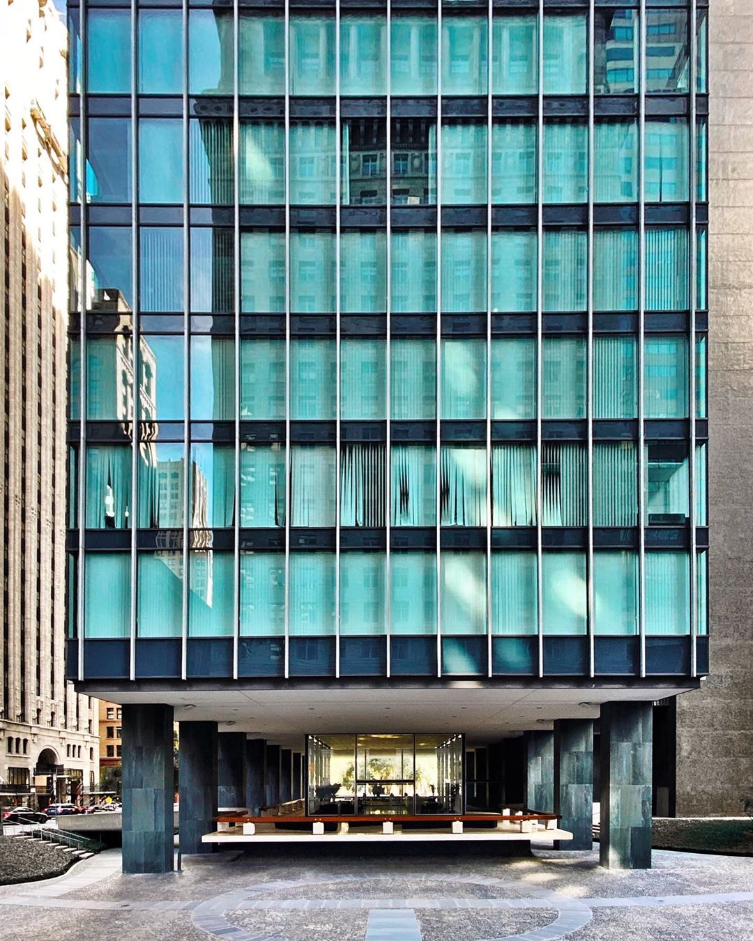 Exterior view of a modern building with a glass facade and a column-supported entrance; a portion of an older building is reflected.