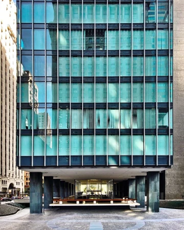 Exterior view of a modern building with a glass facade and a column-supported entrance; a portion of an older building is reflected.