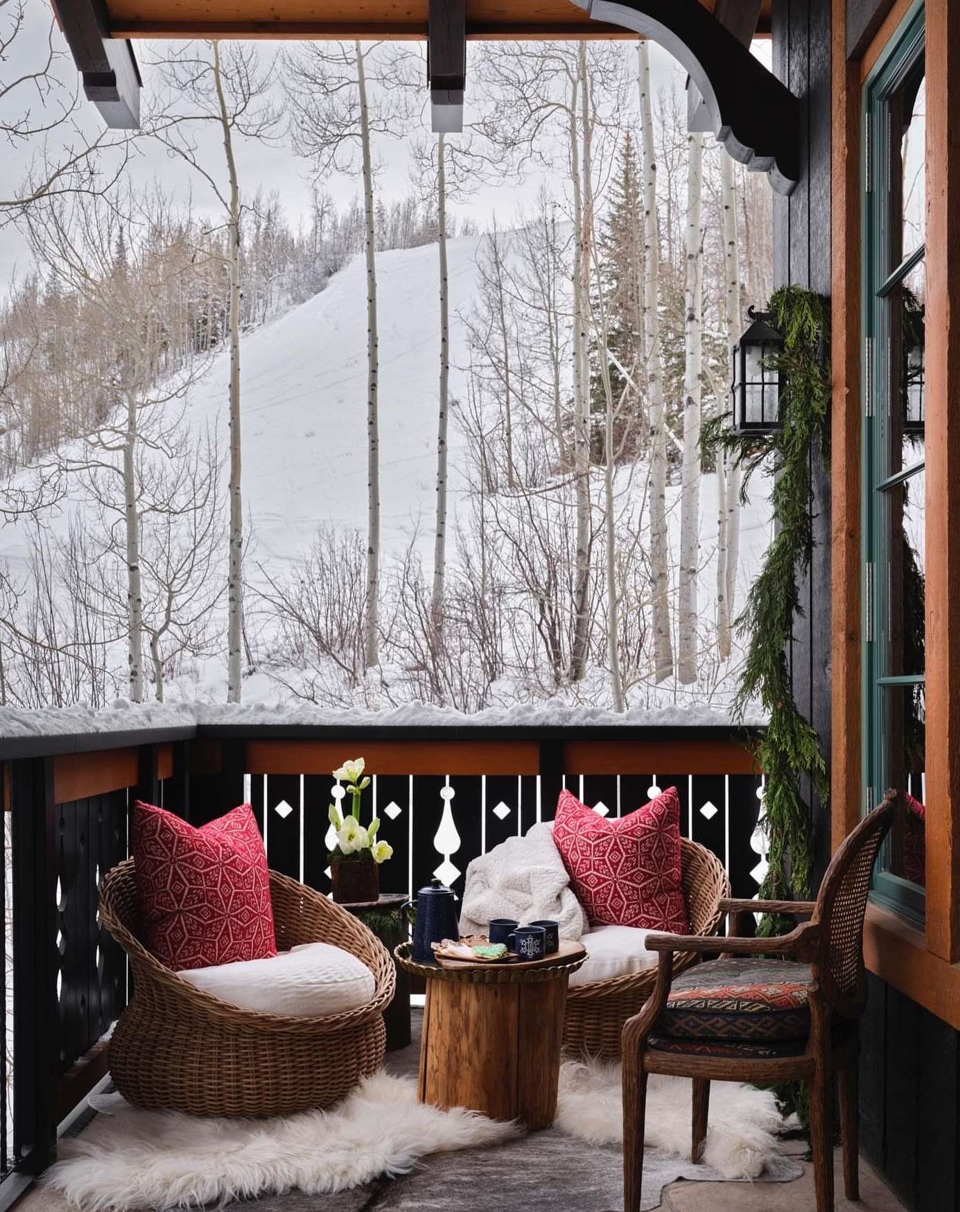 Outdoor seating area with wicker chairs and red cushions on a porch, offering a view of a snowy mountain landscape and bare trees.
