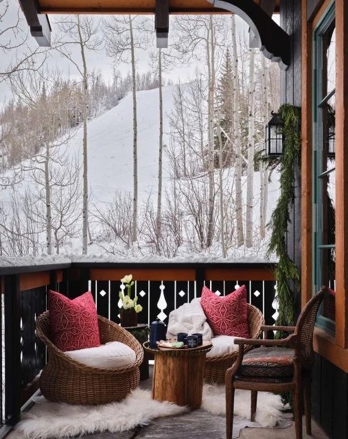 Outdoor seating area with wicker chairs and red cushions on a porch, offering a view of a snowy mountain landscape and bare trees.