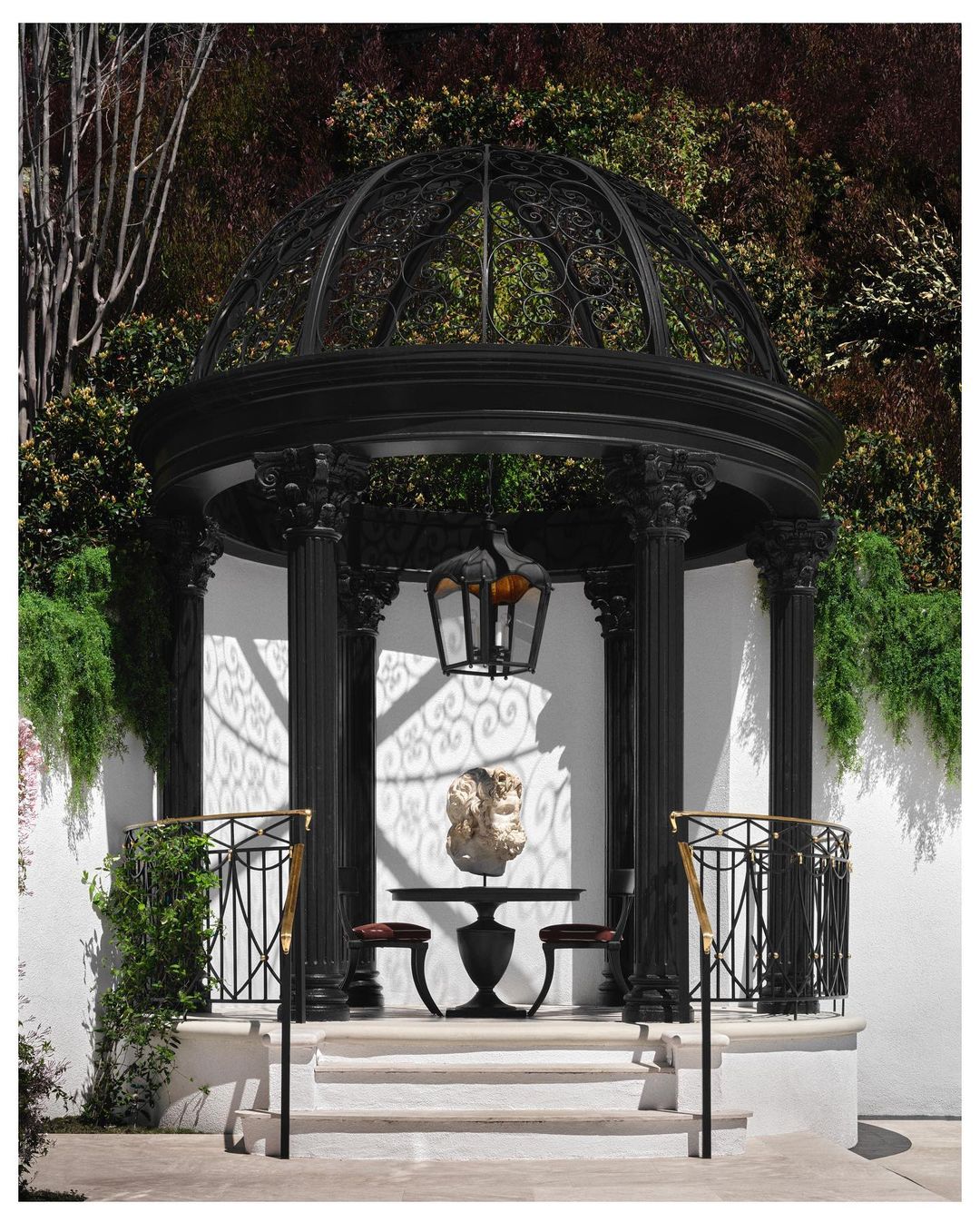 Black metal gazebo featuring a table with a sculpture and two chairs, set on a stone platform against a lush green background.