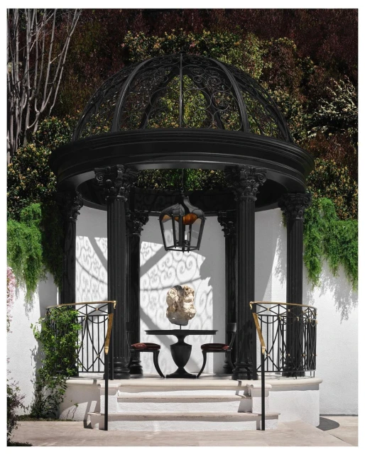 Black metal gazebo featuring a table with a sculpture and two chairs, set on a stone platform against a lush green background.