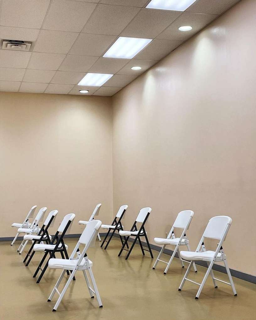 Indoor shot of rows of black and white folding chairs on a beige floor, under fluorescent lights and against a beige wall.