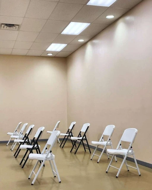 Indoor shot of rows of black and white folding chairs on a beige floor, under fluorescent lights and against a beige wall.