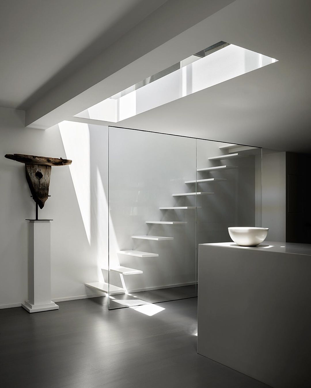 Minimalist interior with floating stairs behind glass, a sculpture on a pedestal, modern white kitchen counter, and light from a skylight.