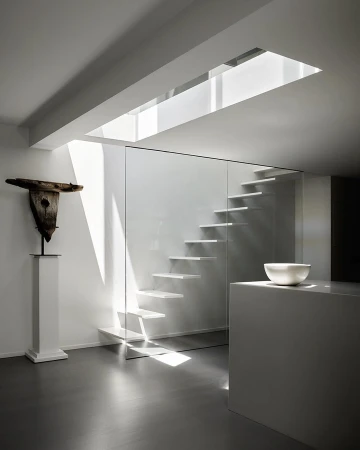 Minimalist interior with floating stairs behind glass, a sculpture on a pedestal, modern white kitchen counter, and light from a skylight.