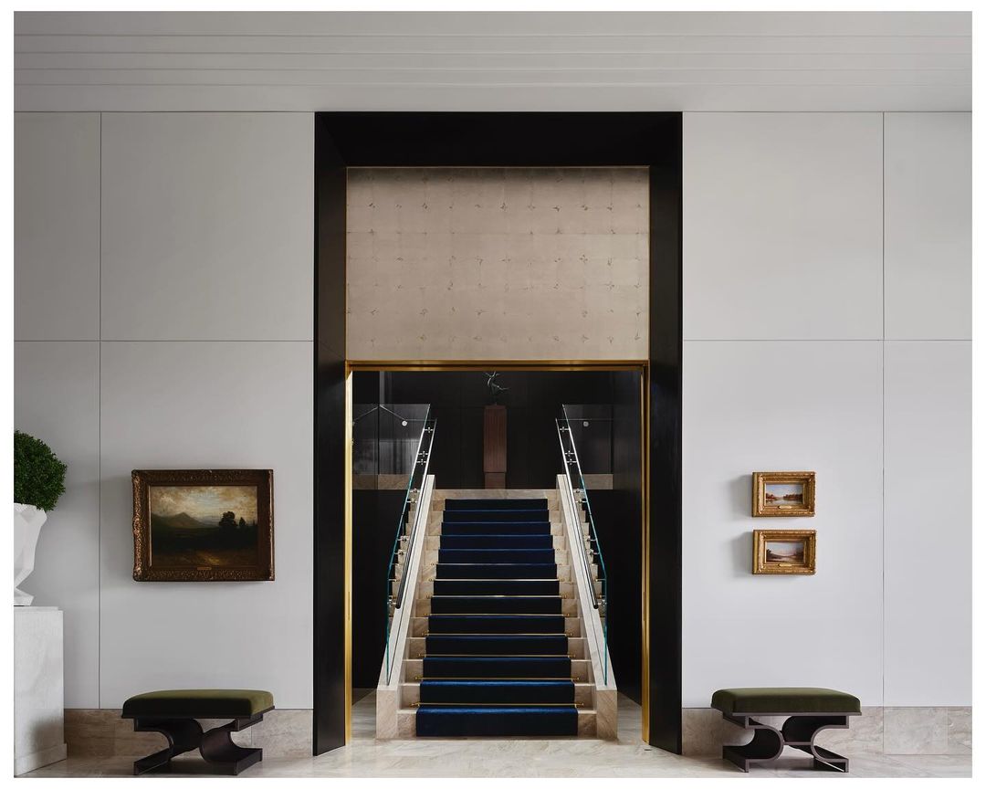 Interior design featuring a staircase with a blue carpet, framed artwork on the wall, and two benches in a bright, modern space.