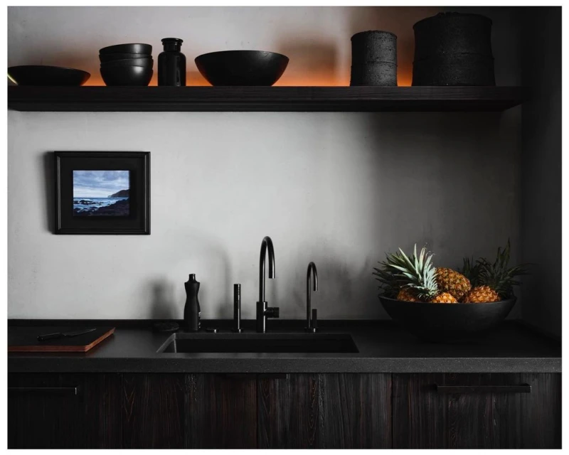 Modern kitchen interior features dark wood cabinets, black hardware, black bowls and utensils on a shelf, pineapple in a bowl, and framed artwork.