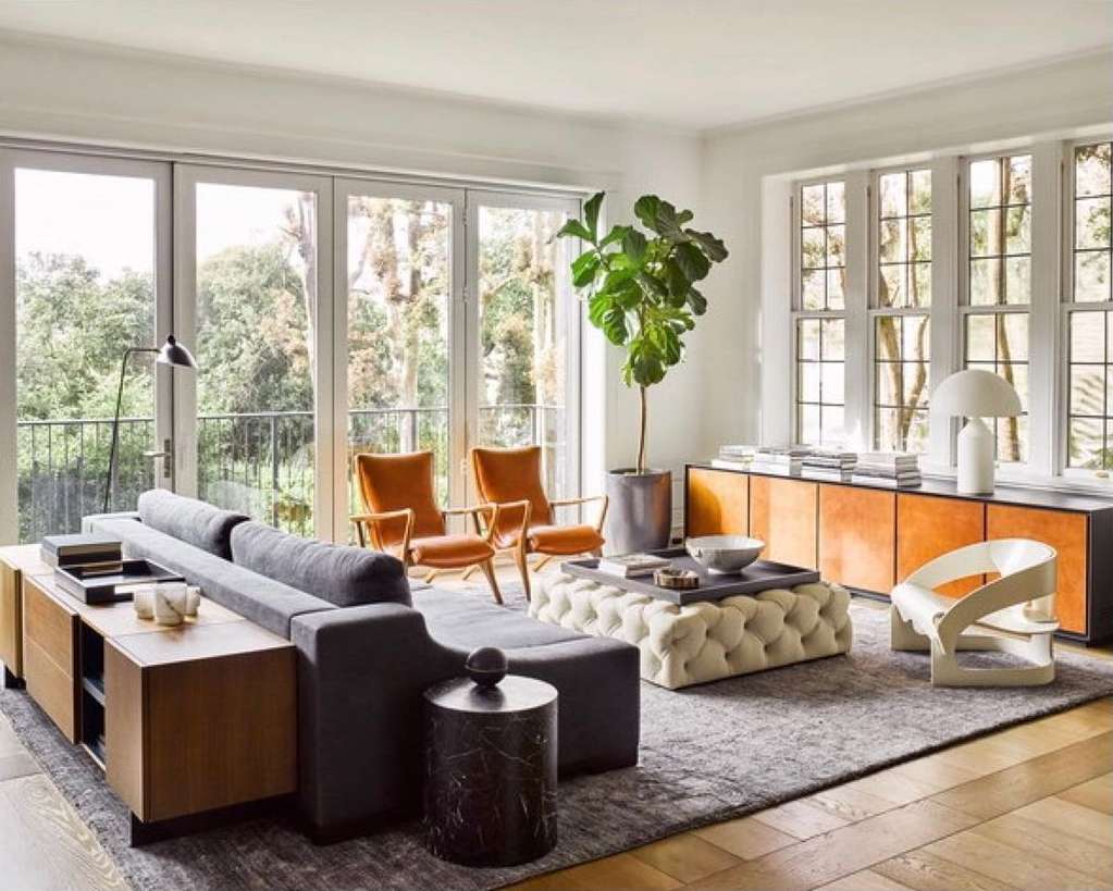 Living room interior with a grey couch, orange chairs, tufted ottoman, wooden cabinet, potted tree, large windows, and natural light.
