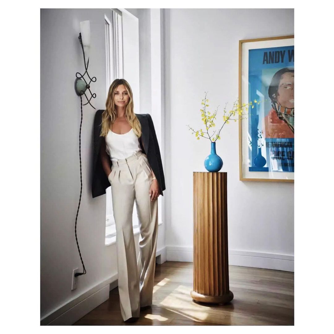 Adult woman in elegant casual wear poses indoors against wall with modern decor, natural light, wooden accent, and artwork.