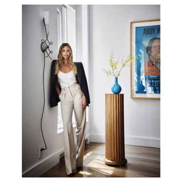 Adult woman in elegant casual wear poses indoors against wall with modern decor, natural light, wooden accent, and artwork.