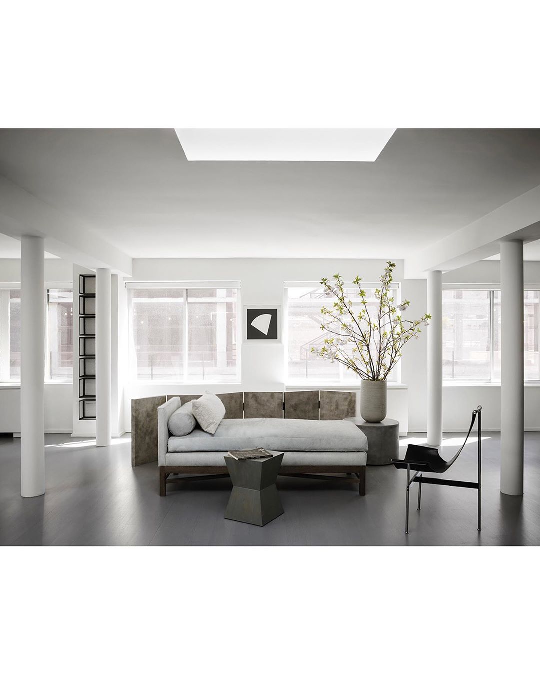 Living room interior with a long daybed, geometric table, art print, columnar supports, large windows, and a vase of leafy branches.