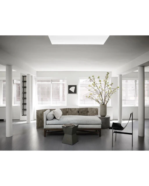 Living room interior with a long daybed, geometric table, art print, columnar supports, large windows, and a vase of leafy branches.