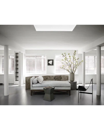 Living room interior with a long daybed, geometric table, art print, columnar supports, large windows, and a vase of leafy branches.