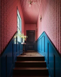 Interior shot of a hallway with pink striped wallpaper, blue paneled wainscoting, and dark wooden stairs leading up to a closed door.