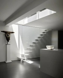 Minimalist interior with floating white stairs, a glass partition, and dramatic natural skylight illumination.