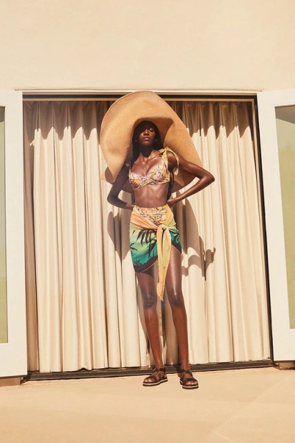 A black woman is posing in a swimsuit and sarong with a giant straw hat, standing in front of a beige wall with a cream curtain.