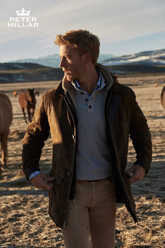 Blonde adult male wearing a brown jacket and khaki pants stands in a field with horses grazing in the background in a rural setting.